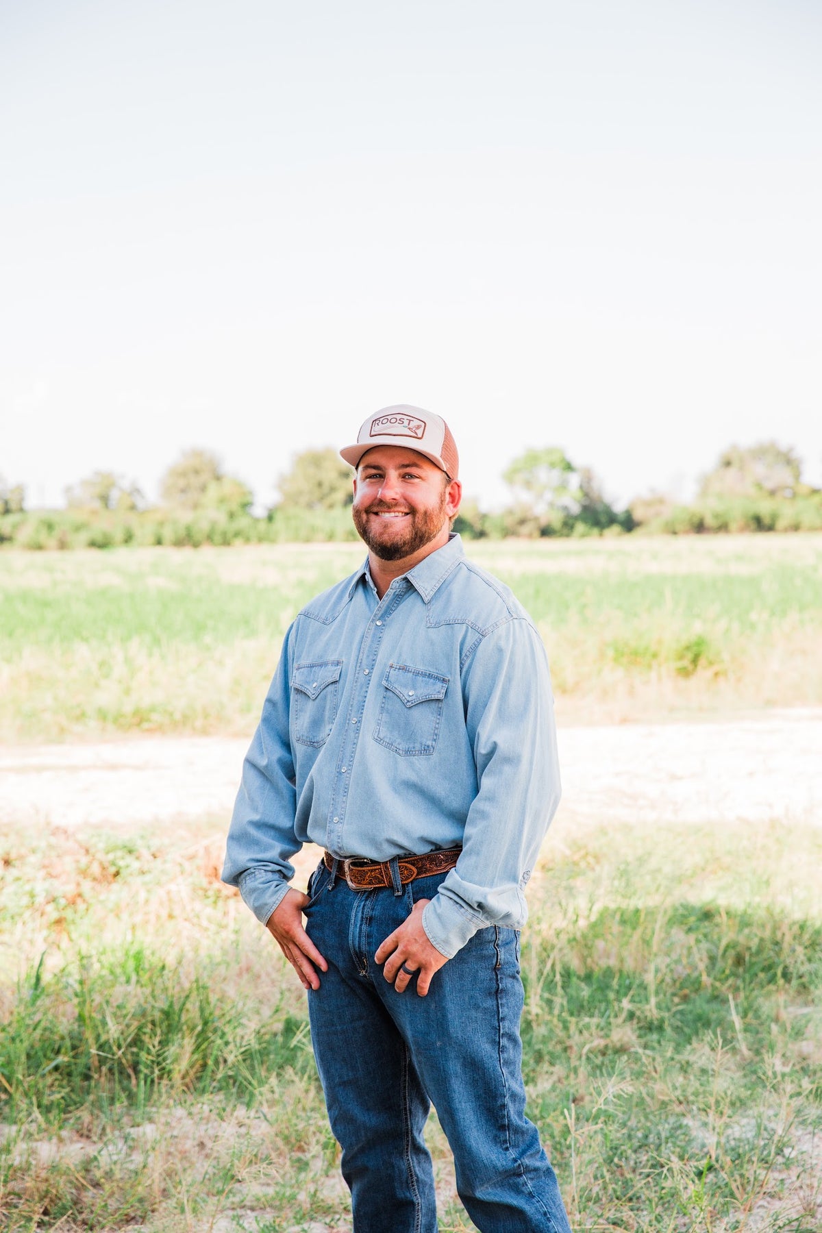 Model facing the camera in a tan Roost Logo Trucker Hat with brown mesh sides, styled with a denim shirt in a grassy field.