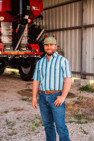 Model wearing the Tristan blue striped short sleeve pearl snap shirt tucked into jeans beside red farm equipment.
