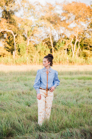 Full-length Western outfit with the chambray Lucky Trails Horseshoe Blouse by Turquoise Haven and cream horse-print pants.