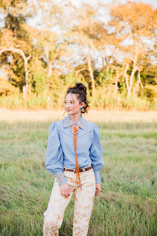 Lucky Trails Horseshoe Blouse by Turquoise Haven in chambray blue, styled with a bolo tie and cream Western-print pants.