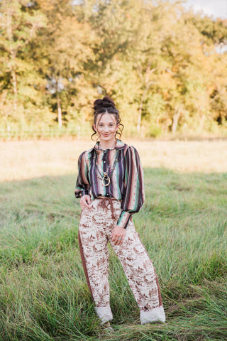 Turquoise Haven Sagebrush Trails Serape Blouse with green and brown stripes, styled with cream ranch-print jeans outdoors.