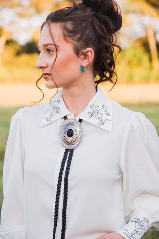 Close-up of bucking horse embroidery on the white pointed collar of the Turquoise Haven Wild Rein blouse, worn with a bolo tie.