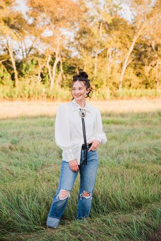 Turquoise Haven Wild Rein Bucking Horse Blouse in white, styled with a bolo tie and distressed blue jeans outdoors.