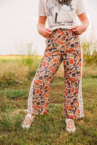 Front view of Varsity Rodeo duck camo pants with wide legs and white lace side trim, styled with a graphic tee and sneakers.