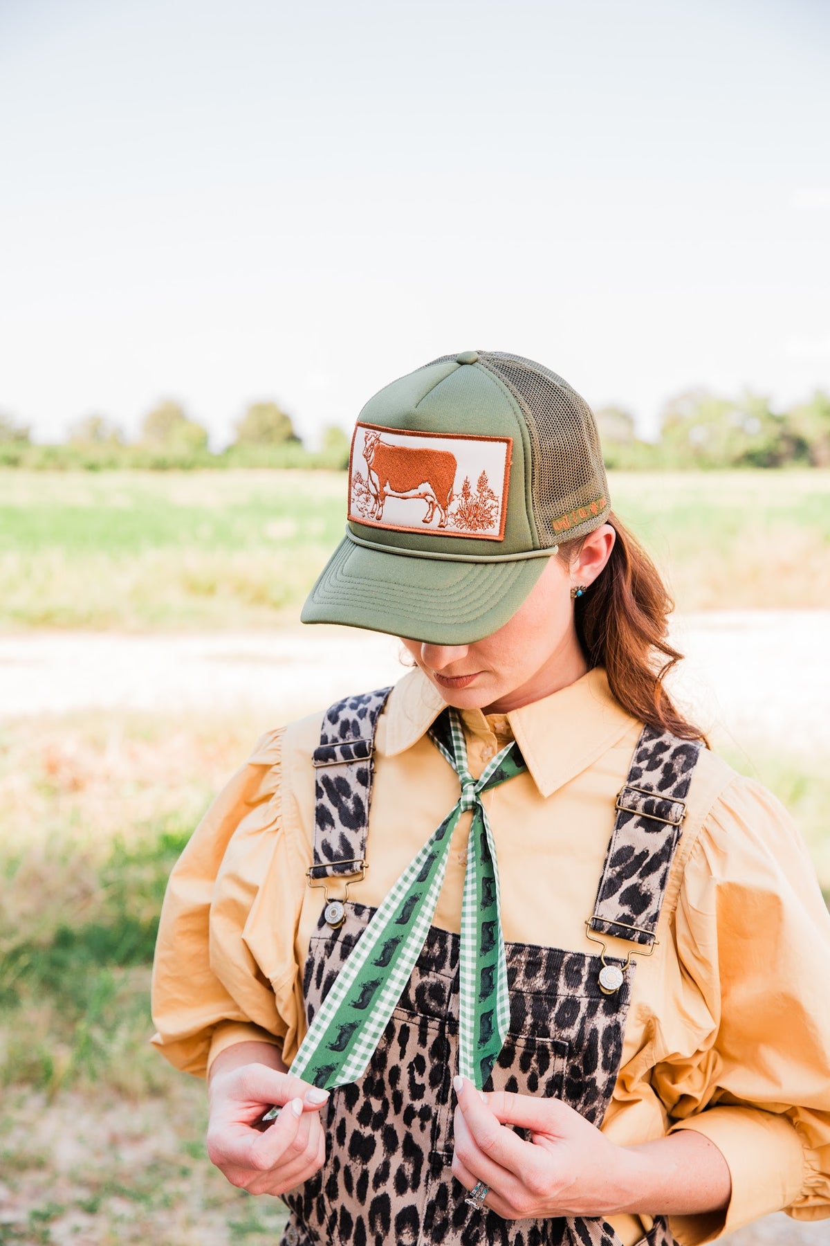 Model wearing a green Vintage Hereford Trucker Hat with a cattle-print neck scarf, tan blouse, and leopard overalls.
