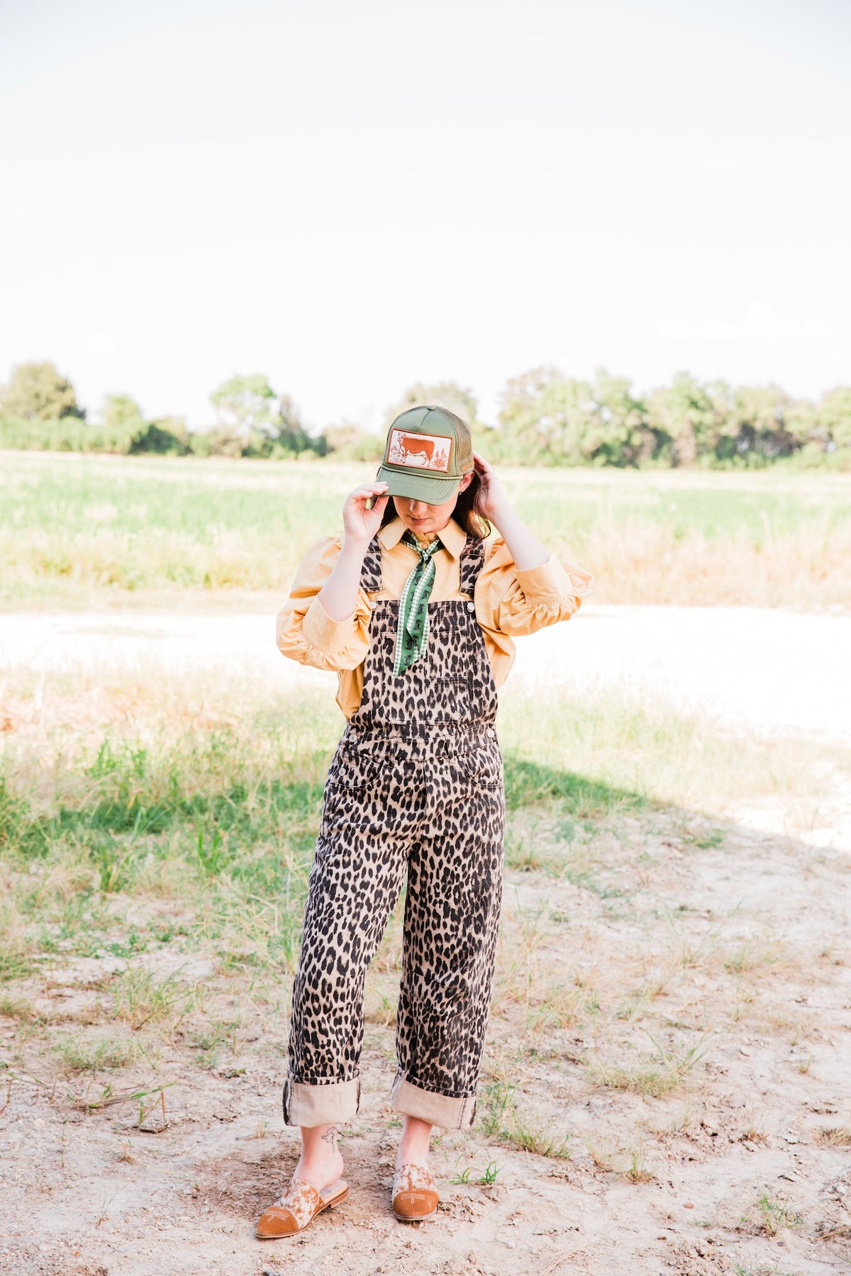 Full-length outfit with the green Vintage Hereford Trucker Hat, leopard overalls, a tan blouse, and a green neck scarf.