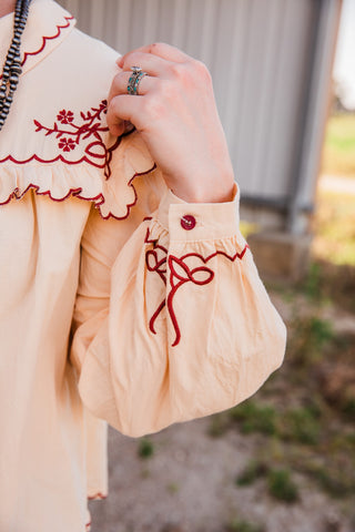 Winchester Top sleeve close-up with burgundy bow embroidery, a matching cuff button and scalloped floral yoke trim.