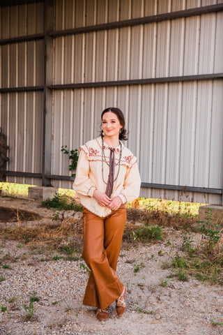 Model wearing the Winchester Top with burgundy floral embroidery and a dark neck ribbon, paired with tan pants and boots.