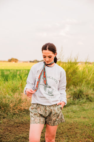 Front view of women’s camo boxer shorts with a woodland branch-and-leaf print, styled with a gray sweatshirt and neck scarf.