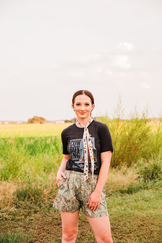 Model wearing women’s camo boxer shorts with a black cowboy graphic tee and a cream printed neck scarf in a grassy field.
