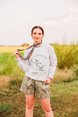 Women’s woodland camo boxer shorts styled with a gray duck graphic sweatshirt and a colorful neck scarf outdoors.