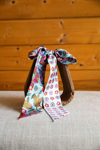 Cottage Garden silk twilly by XOXO Art & Co tied in a bow, with multicolor flowers on soft blue fabric and pointed ends.
