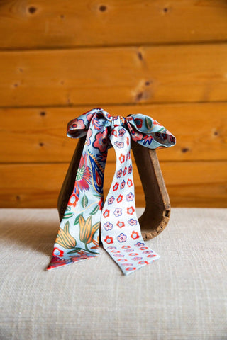 XOXO Art & Co Cottage Garden silk twilly displayed on a wooden stirrup, showing large colorful blooms and small floral motifs.