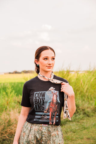 Cowboying black graphic tee by XOXO Art & Co with stacked lettering and a cowboy on a brown horse, styled with a neck scarf.