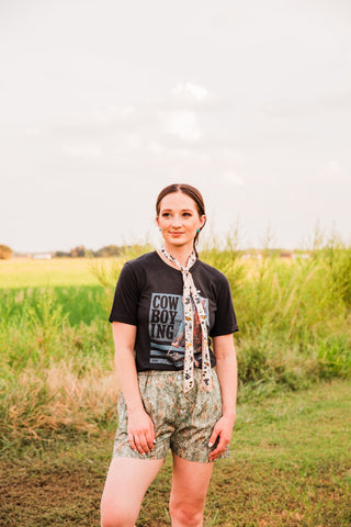 Cowboying Graphic Tee by XOXO Art & Co on a model in a grassy field, paired with camo boxer shorts and a cream neck tie.