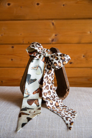 XOXO Art & Co Wild Horses silk twilly with pointed tails displaying horse-and-horseshoe artwork beside brown leopard spots.